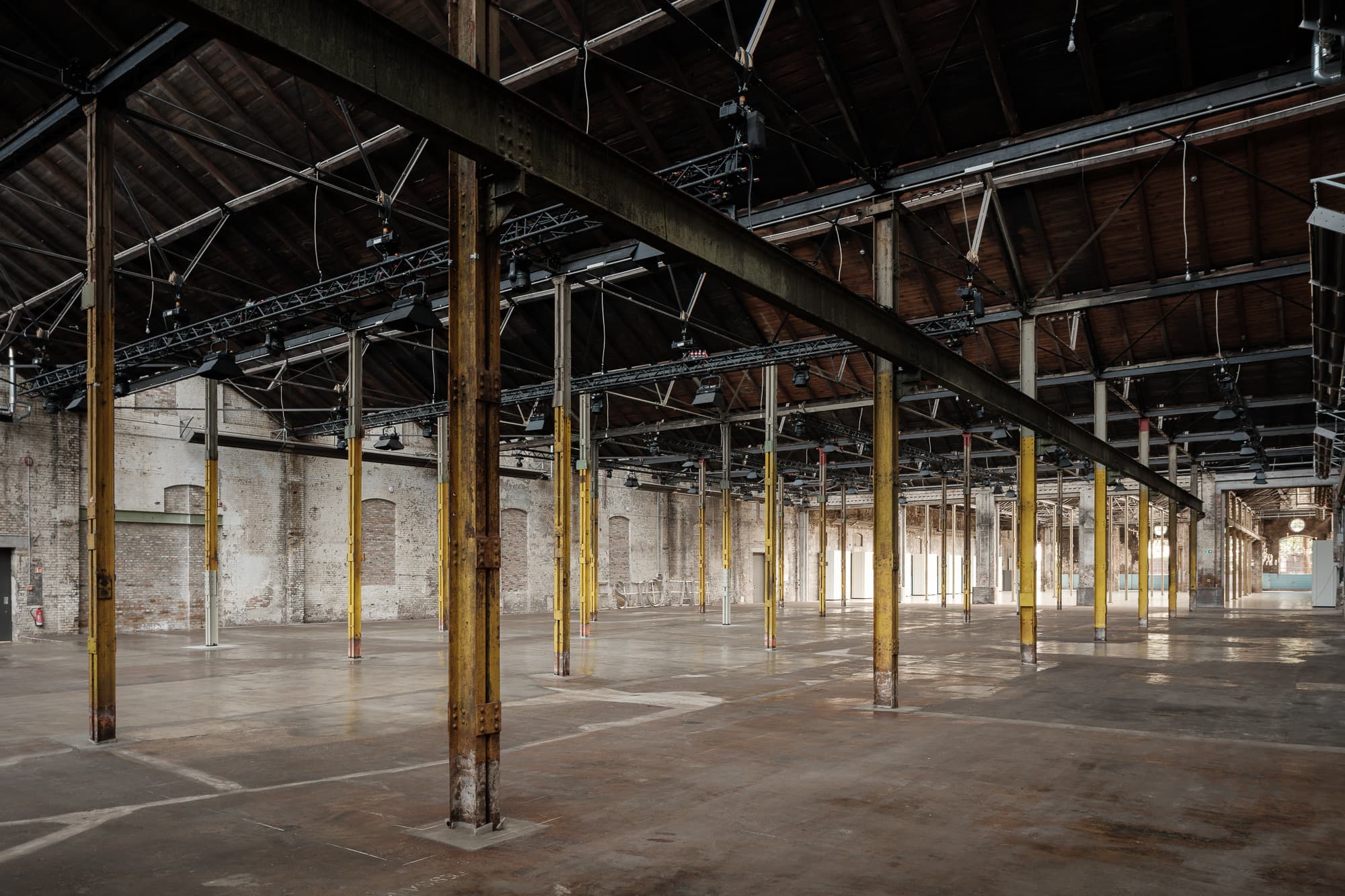 Inside Wilhelm Hallen: a long industrial hall with yellow steel columns, a timber roof and a concrete floor